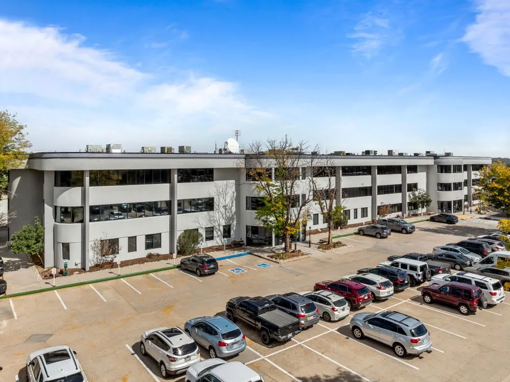 Large gray office complex with multiple levels, full parking lot, trees, and clear blue sky