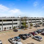 Large gray office complex with multiple levels, full parking lot, trees, and clear blue sky
