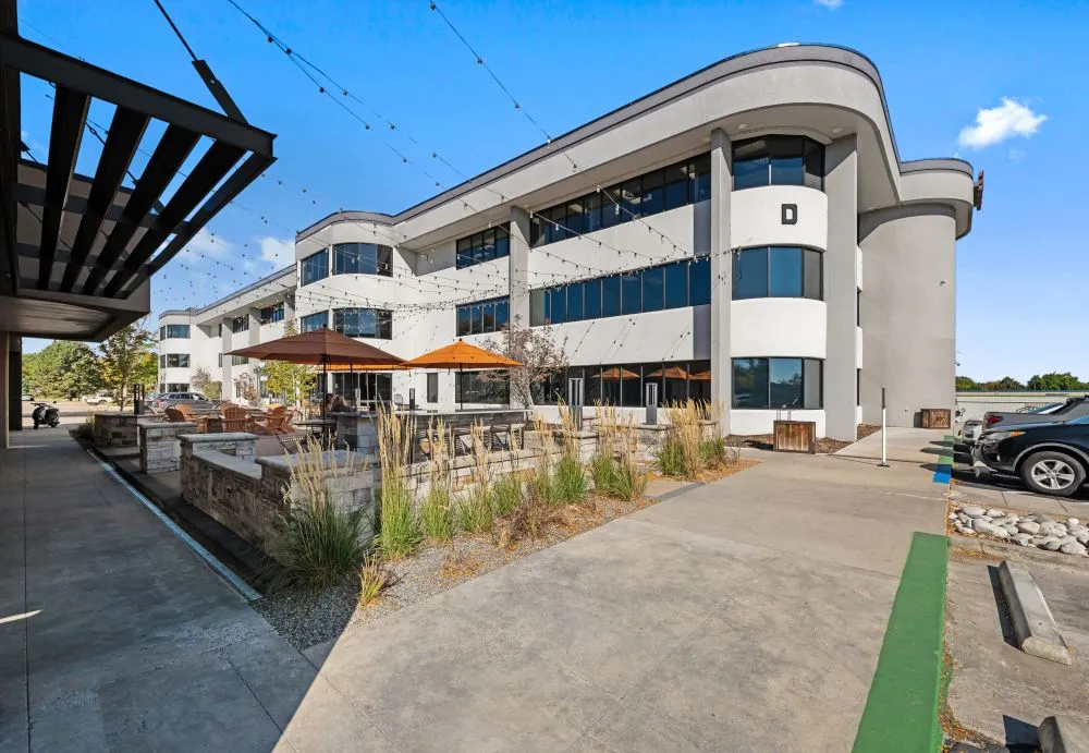 Modern multi-story office building with curved white facade, outdoor seating, and covered walkway under blue sky