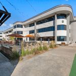 Modern multi-story office building with curved white facade, outdoor seating, and covered walkway under blue sky