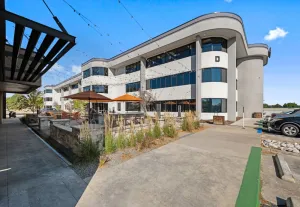 Modern multi-story office building with curved white facade, outdoor seating, and covered walkway under blue sky